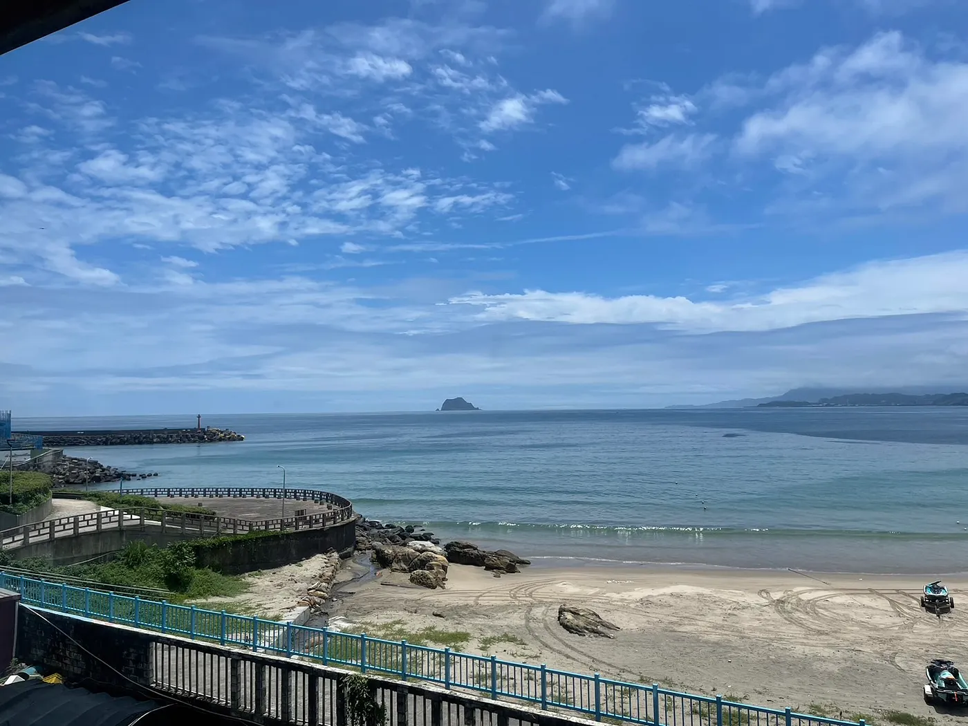 晴天海岸風景,沙灘與防波堤延伸至遠方海面,遠處可見一座小島,天空湛藍、白雲飄散。