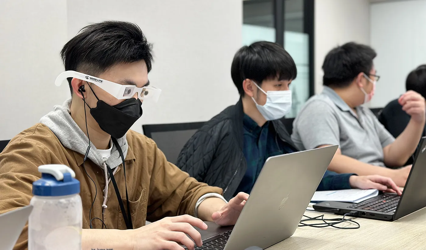 三位團隊成員正在電腦前工作，畫面中的第一位同事帶著特殊眼鏡，模擬視障者所看到的畫面。
